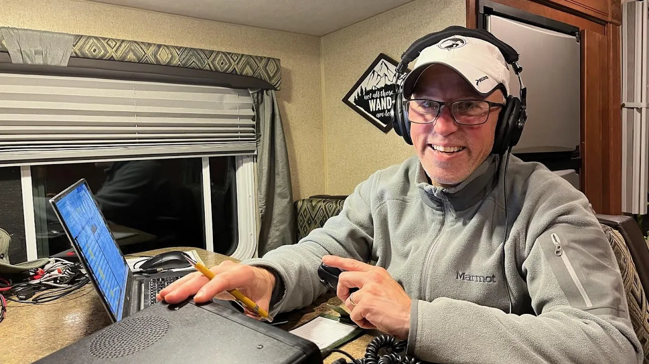 A smiling operator in a ball cap and headset logging contacts on a laptop and radio at a table inside an RV.