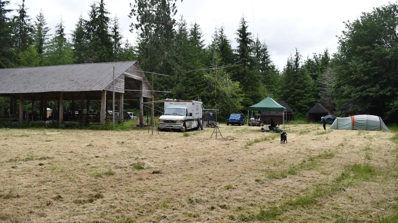 MicroHAMS Field Day at Camp Freeman — a large covered pavilion, the Snohomish County communications van, antenna masts, pop-up canopies, and tents spread across a grassy clearing ringed by tall evergreens.