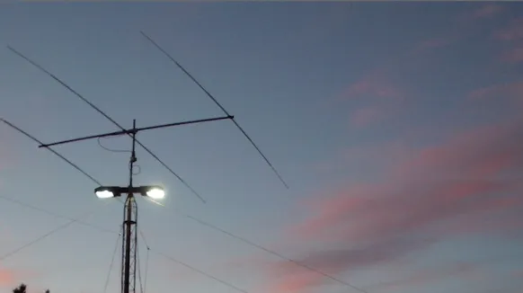A tribander beam antenna silhouetted against a pink-and-blue sunset sky, with a work light glowing below the boom, atop the building 40/41 parking garage.