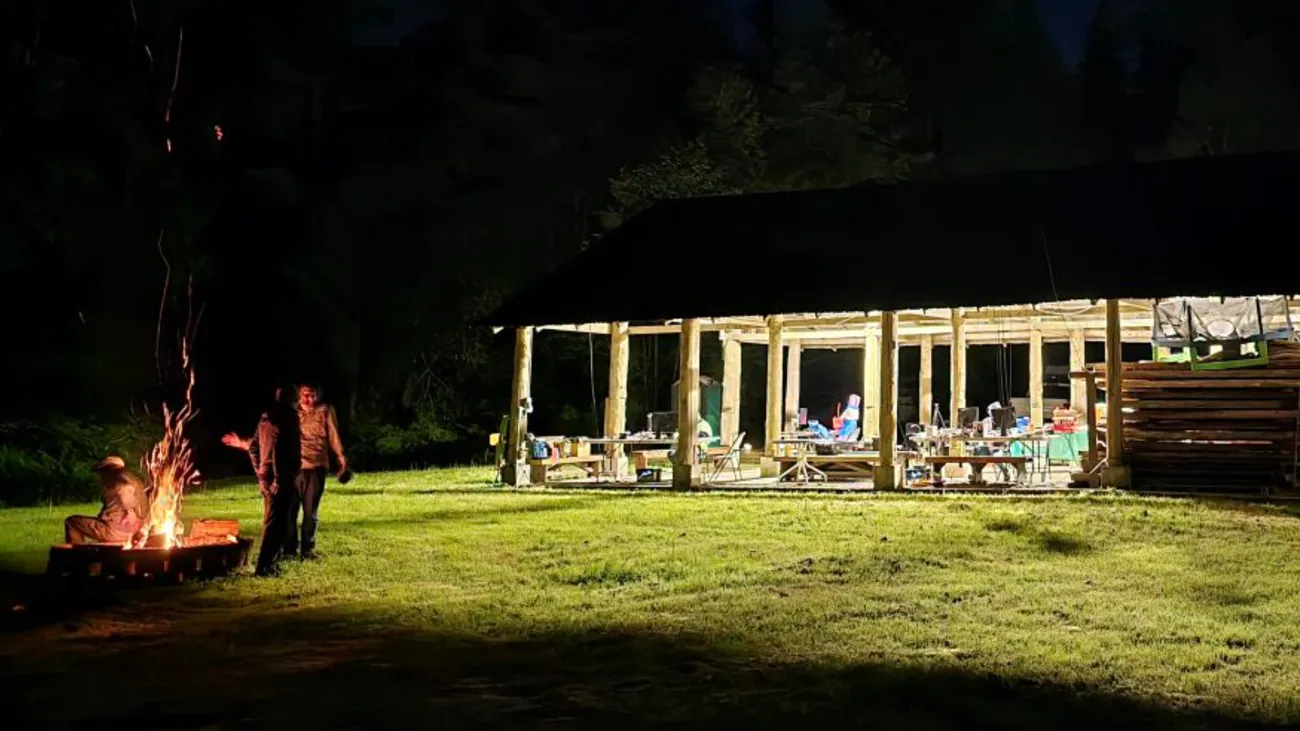 N7OS Field Day at night — the Camp Freeman pavilion lit from within with operating stations, a campfire and two operators at left, and the Snohomish County Emergency Management communications van at right beneath a VHF antenna.