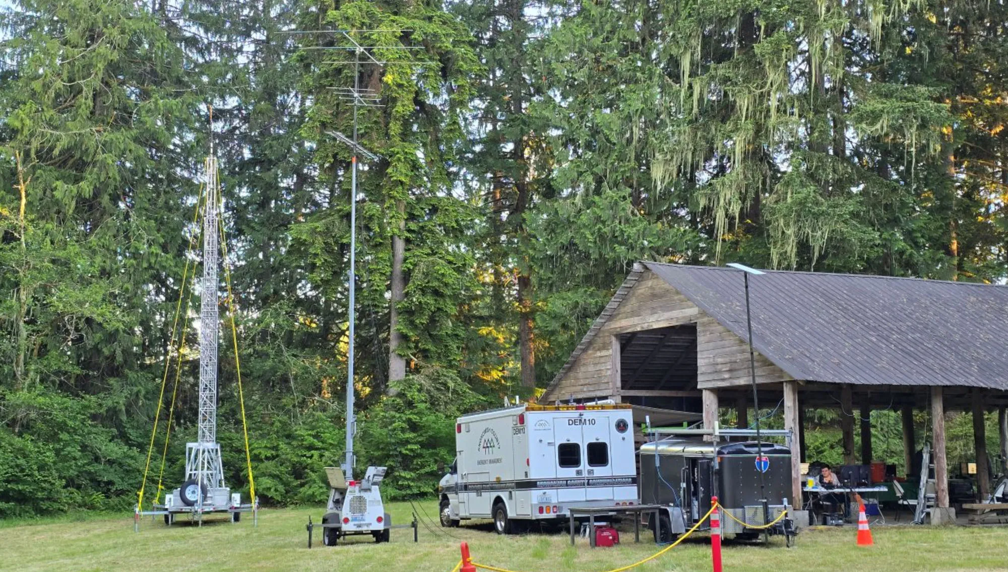 The N7OS VHF station at Camp Freeman — a mast trailer with stacked VHF/UHF Yagis and a solar light tower on the left, the Snohomish County DEM communications van (DEM10) in the center, and an operator working from the covered pavilion at right.